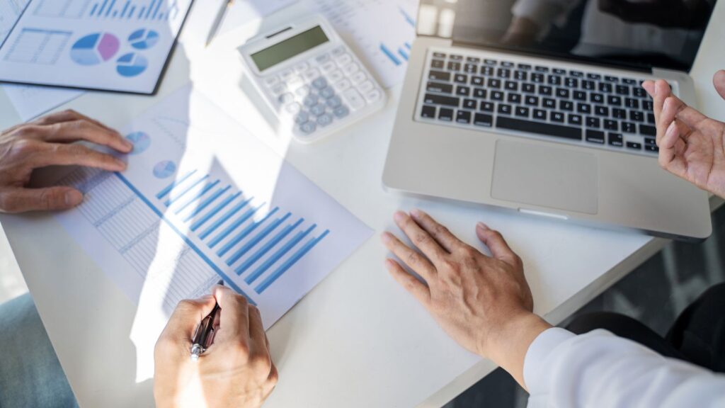 Business startup team analyzing printed charts and graphs around a laptop, discussing financial data and performance metrics during a strategy meeting.