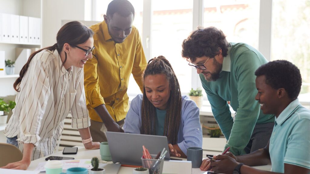 Diverse business team collaborating around a laptop in a bright modern office, representing teamwork and improved collaboration—one of the key benefits of Business Central SaaS.