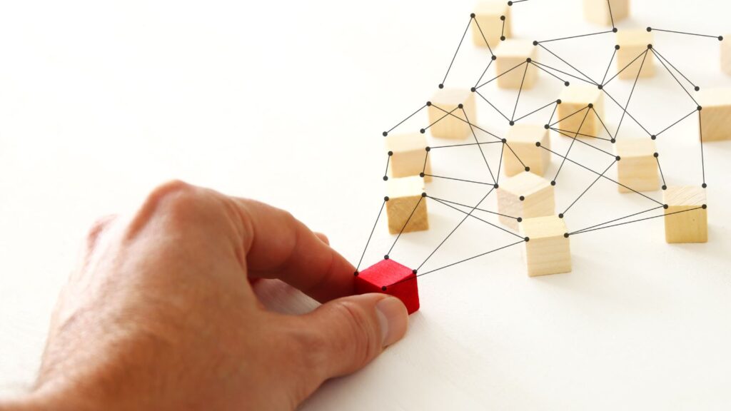 Hand connecting red wooden block to network of linked cubes, representing multi-entity management and organizational connectivity.