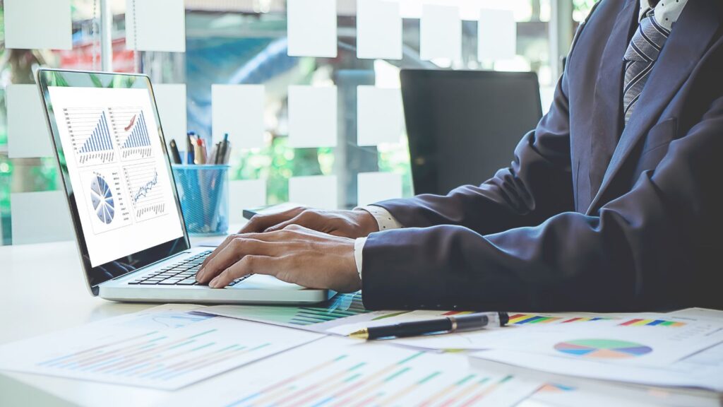 A business professional in a suit working on a laptop displaying charts and graphs, with printed financial reports and a pen on the desk, representing data analysis, business strategy, or financial planning.
